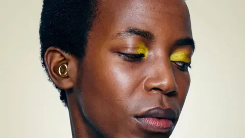 Loop A close-up of someone wearing yellow eye shadow. In their ear is a gold loop-style ear-plug.