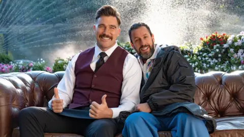 Scott Yamano/Netflix Travis Kelce with both thumbs up sitting on a leather sofa with Adam Sandler, both smiling, on the set of Happy Gilmore 2