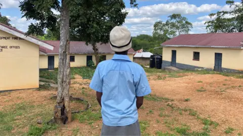 Wycliffe Muia / BBC A student wearing a light blue short-sleeved school shirt, a grey skirt, and a cream-colored knit hat stands beneath a large tree in the courtyard of Manooni Primary School in Kenya