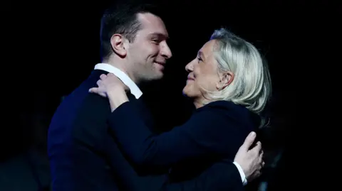 Getty Images French far-right Rassemblement National RN party's President Jordan Bardella and the President of the party's parliamentary group Marine Le Pen greet each other during a party rally following the results of the first round of France's 2026 municipal elections, in Chalons-en-Champagne. They are smiling and embracing. 