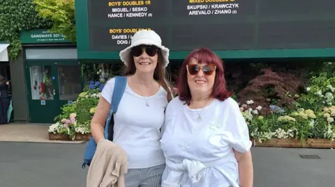 Supplied Sherly and Marianne are standing in front of a fixtures board in the Wimbledon grounds