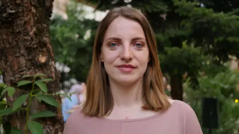 Marjana Koceku is smiling directly at the camera. She has dark blonde hair in a long bob and is wearing pink top. Behind her are some trees. 