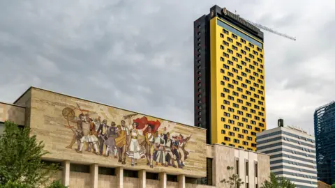 Getty Images  Tirana's central Skenderbeg Square. In the foreground is a large mural from Albania's communist era in the socialist-realist style. Behind this building there are cranes and skyscrapers under construction. 
