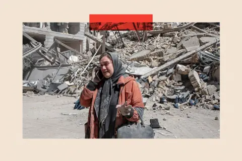 Getty Images A woman in an orange jacket and grey headscarf talks on the phone in front of a destroyed building