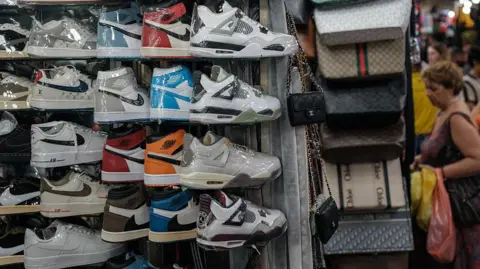 Getty Images Plastic-wrapped Nike sneakers racked at a marketplace alongside handbags while people shop in the background