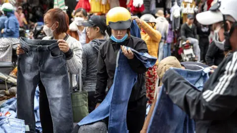 Getty Images Women, including one wearing a bright yellow motorcycle helmet, look at pairs of jeans at an outdoor market