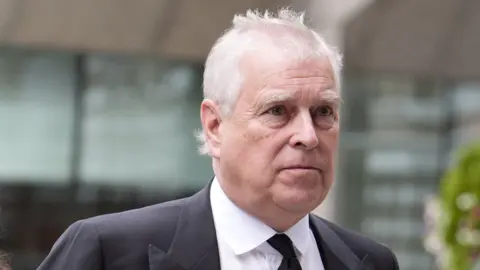 PA Wire/PA Images Close-up portrait of a person with short white hair wearing a dark suit jacket, white shirt and black tie. The person is positioned in the foreground and facing slightly to the side. The background is out of focus, showing glass windows, a light-coloured building structure and greenery. No bags, bins, signs or markers are visible.
