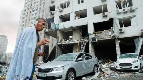 EPA/Shutterstock Injured man wearing a blue blanket standing in front of an apartment building that has been badly damaged