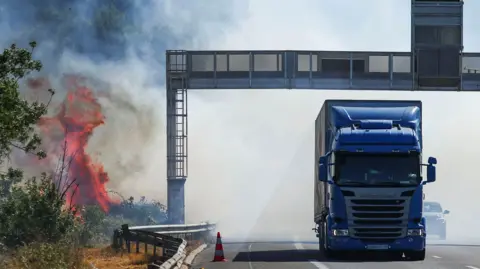 REUTERS/Abdul Saboor A truck drives next to a fire as a firefighting plane (not pictured) flies to battle wildfire near the A9 motorway near Beziers, France, July 3, 2026. 