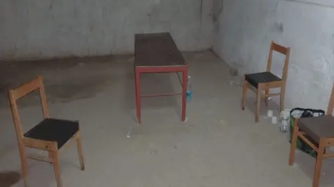 Telegram A bare room with concrete walls and floor. In it are three wooden chairs and a small wooden table, with a few plastic water bottles on the floor.