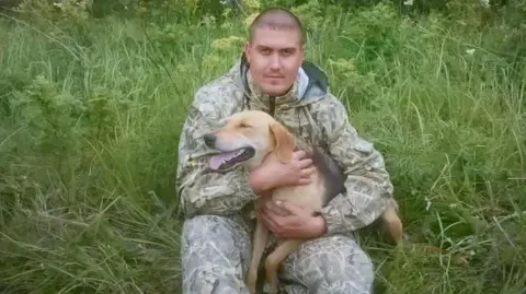 OK.ru Andrey Spivak sitting in long grass holding a beige and black dog. He is wearing a camouflage-style outdoor suit, has hair shaved closed to his head and is looking directly at the camera.