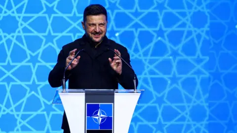 Reuters President Volodymyr Zelensky is dressed in a black military jacket and trousers, speaking behind a podium at Nato. He gesticulates whilst he talks into the microphones. Behind him is a blue background with stars on it.