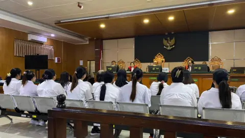 BBC Indonesian Two rows of people dressed in white sit on benches in a court room, facing a panel of judges