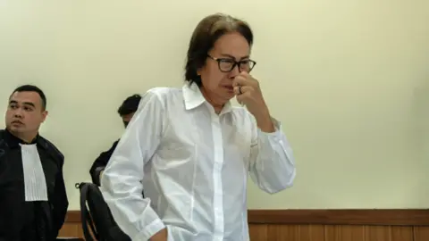 BBC Indonesian A woman wearing a white shirt and black-rimmed glasses looks emotional and holds a tissue up to her nose