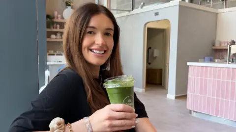 Jess Bloom Jess smiles, holding an iced matcha latte in a cafe. She has long brown hair and wears a black top