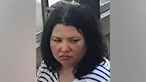 Interpol A woman with dark hair and a serious expression looks down. She wears a striped black and white shirt.