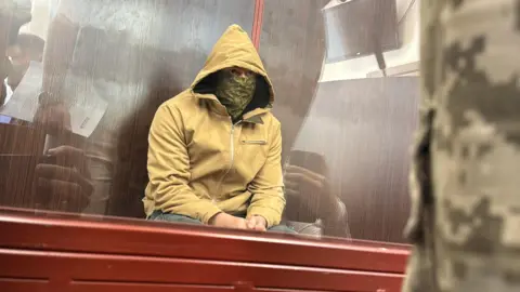 A man sits with a hood on and yellow jacket and face mask