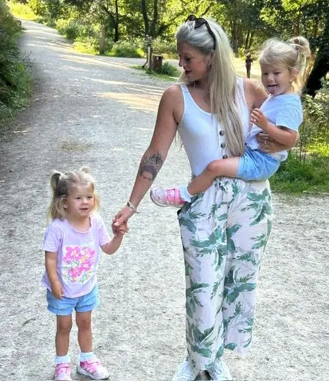 Brioney Garrett Brioney carries one twin while holding the other's hand while walking along a grey, stoney path bordered by greenery and trees in the sunshine - they all have blond hair