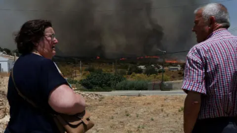 Reuters A concerned looking woman with a dark brown bob, wearing a t-shirt, looks at a greying man who is wearing a checked shirt. In the distance, dark smoke is seen billowing above a line of fire going through shrubland.