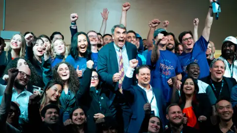 A group of young people cheer and pump their fists in the air. In the middle, an older man in a grey suit and a purple and yellow tie, pumps his fist and shouts.