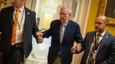 Getty Images Mitch McConnell, wearing reading glasses and a blue suit jacket, leans on the arms of two men also wearing suits 