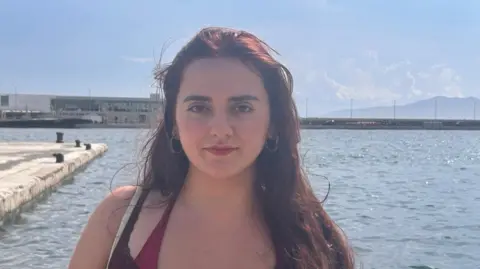 Héloïse H The image is of a young woman with long, dark brown hair falling over her left shoulder. She is looking directly into the camera with a calm expression. She has dark eyebrows, dark eyes, and is wearing a brown top over a burgandy swimsuit, and has small, metallic hoop earrings. Behind her is a wide expanse of calm water with a concrete pier jutting out and several bollards along the edge. In the distance is a long, flat-roofed building and beyond that is a low mountain. 