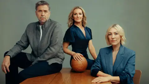 Match of the Day presenters Mark Chapman, Gabby Logan and Kelly Cates pictured sitting at a desk, holding a ball