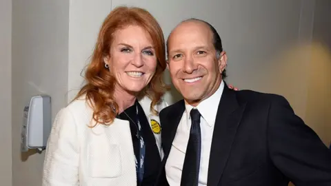 Getty Images Sarah Ferguson and Howard Lutnick pose together indoors in front of a light-colored wall. One person wears a white textured jacket over a dark top with a lanyard and a round yellow badge, while the other wears a dark suit, white shirt and tie. Both face the camera. A wall-mounted dispenser is visible on the left side of the image.