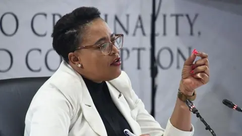 Gallo Images via Getty Images Brig Rachel Matjeng dressed in a white jacket and dark top underneath speaks into a microphone. Behind her, out of focus, is the banner for the Madlanga Commission.