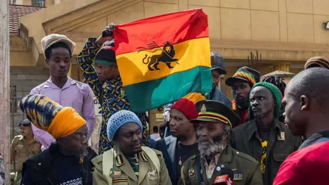 AFP via Getty Images Rastafarians gathering outside a Nairobi court as a bearded man speaks to reporters, while a Lion of Judah flag is held aloft behind him 