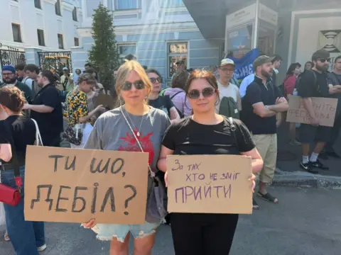 Two young protesters hold up signs reading "What are you doing, you idiot?" and "This is for all of those who couldn't be here"