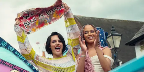 Netflix Still from Heartstopper Forever showing a Pride march with cast members Bel Priestley and Yasmin Finney.