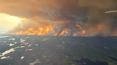 Government of Manitoba /Handout/Getty Images A photo taken from a plane showing green forest interspersed with bodies of water. In the distance is a fire covering the whole horizon and thick black billowing smoke