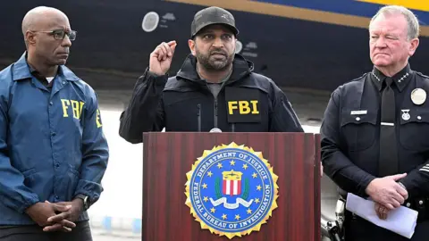 Getty Images Kash Patel wearing an FBI jacket speaking at a podium with an FBI agent standing to his right and a police officer to his left.