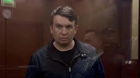 Reuters A darkened photo of a man in court wearing a dark jacket, standing behind a glass screen