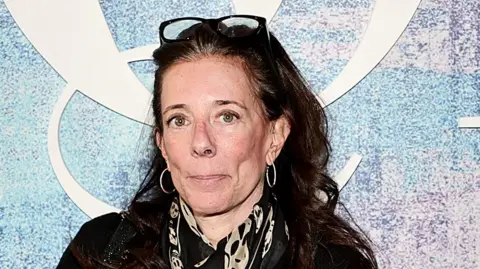 Getty Images Faith Kates, a woman with long brown fair, glasses perched on top of her head, wearing black with a gold-and-black silk scarf, poses for the cameras at a Business of Fashion event with part of the "BOF" logo visible on a hoarding behind her.