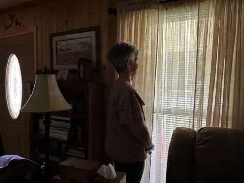 Brandon Drenon/BBC Karen Bowden stands for a portrait in her living room, staring out the window.