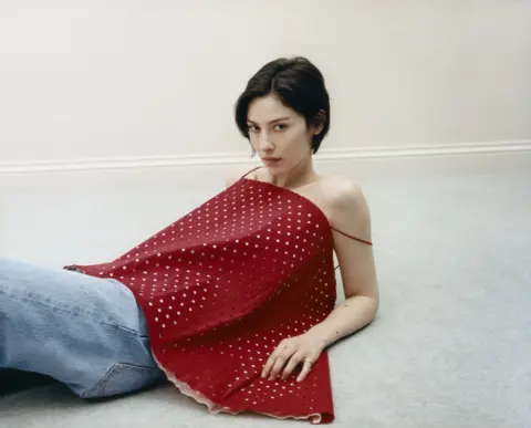 Julie Greve Gracie Abrams in a promotion photo for her third album, Daughter from Hell. She is lying down, propped up on her elbows, in a plain room; wearing a strappy polka dotted top.