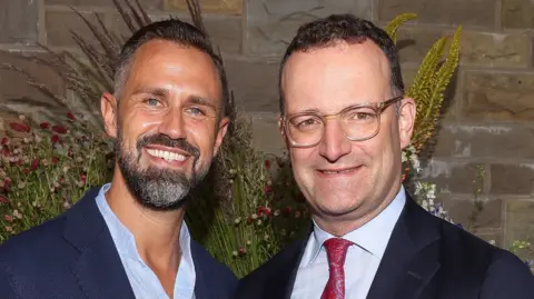 Getty Images Jens Spahn (R) in glasses, a dark jacket and white shirt and red tie beside his husband Daniel Funke who has as beard and a navy jacket with a blue shirt 