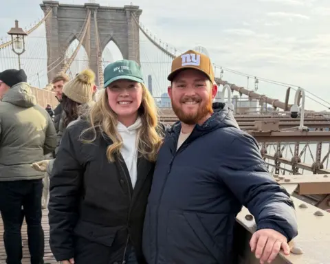 Hannah Leonard-Ripley A woman and a man stand on a bridge in New York, smiling at the camera. They are both wearing coats and baseball caps