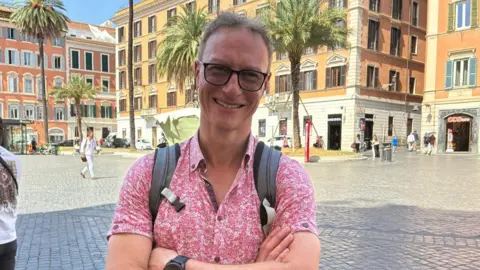 A man with short light brown hair and dark-rimmed glasses, wearing a pink flower patterned shirt and a rucksack, smiling. His arms are crossed. He's standing in the cobbled Piazza di Spagna in Rome.