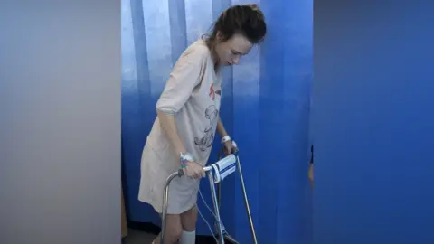 Monica Thomas A woman in hospital using a walker. She has dark hair that has been tied up and she is wearing a long pink nightgown. There are various tubes connected to her and hospital machinery. She is walking in front of a blue curtain that separates hospital beds. 