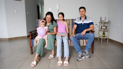 JORGE PEREZ / BBC Camila pictured with her family who are sitting on a sofa in a living room with a tiled floor. She is wearing a pink dress and hair band and is holding a purple balloon. She is sitting on the knee of her mum who has long dark hair and glasses, and is wearing a green jumpsuit. Her sister is in the middle with a purple off-the-shoulder top and jeans, next to her dad who has short dark hair, a blue polo t-shirt and jeans.