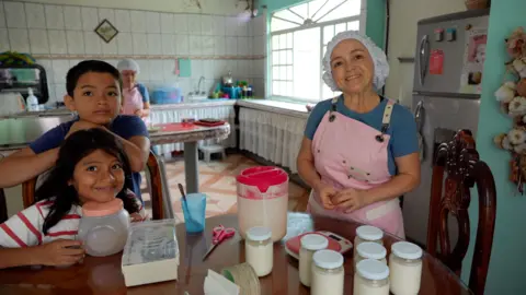 JORGE PEREZ / BBC María Luisa, pictured with her and her sister's children. She is at the right of the frame, smiling, with a pink apron and hair net. The little girl smiles at the camera while sitting on a chair, she has long dark hair, while her cousin leans on the back of the chair. He has short dark hair. María del Cisne is working in the background. 
 