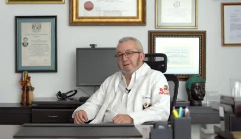 JORGE PEREZ / BBC Dr Guevara in his office, he is sitting at a desk with a wall full of framed certificates. He has short grey hair and beard, with glasses and is wearing a white doctor's coat and white shirt.