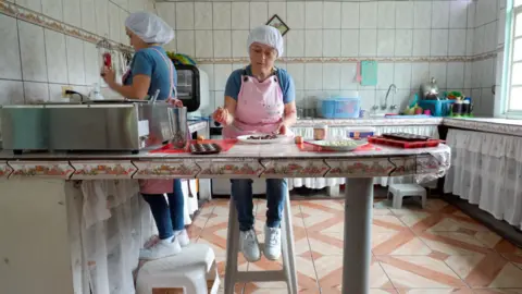 JORGE PEREZ / BBC Maria Luisa is pictured working at a kitchen counter top, sitting on a high stool. She has a pink apron on and a hair net. Her sister works behind her, standing on a step to meet the counter