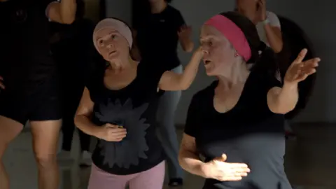 JORGE PEREZ / BBC  María Luisa and María del Cisne at a dance class - they both hold one hand across their stomachs with the other in the air. They both wear black t-shirts and pink hair bands