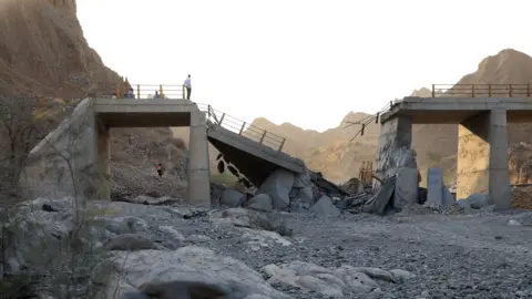 EPA/Shutterstock A damaged bridge with people standing on the remaining structure with mountains in the background, and rubble and rocks in the foreground.