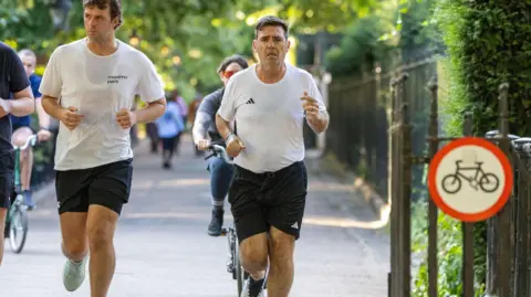 Tayfun Salci Andy Burnham, wearing a white Adidas top and black running shorts, running along a pavement in Green Park