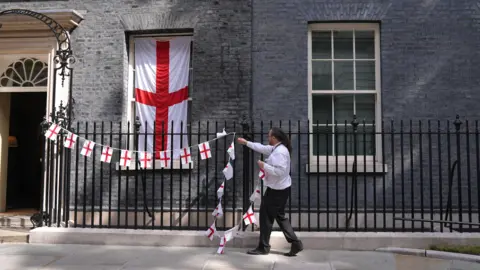 PA Media A man in a shirt and black trousers takes down England flag bunting that was on the railings outside No 10 Downing Street. A large England flag still remains draped in front one of the windows.